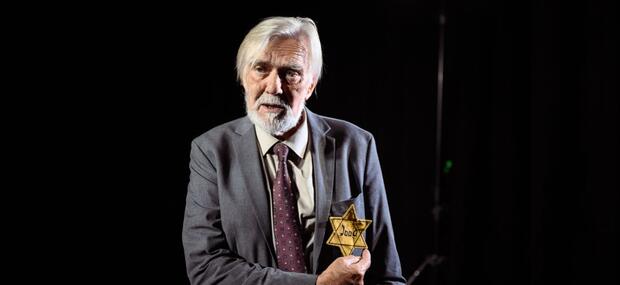 En ældre herre står i jakkesæt med slips og peger på en jødestjerne syet på jakken