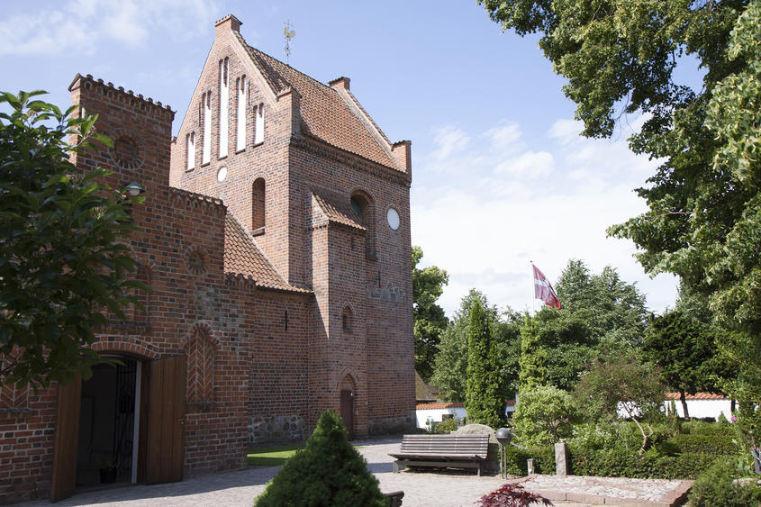 Farum Kirke ligger midt på den gamle kirkegård. Døren står åben og flaget vejrer i baggrunden.