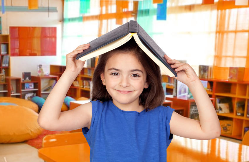 Mørkhåret pige i blå t-shirt, står i forgrunden og holder en bog over hendes hovede som en slags tag. I baggrunden kan man se reoler med bøger og møbler på et bibliotek. Solen skinner ind ad vinduerne.