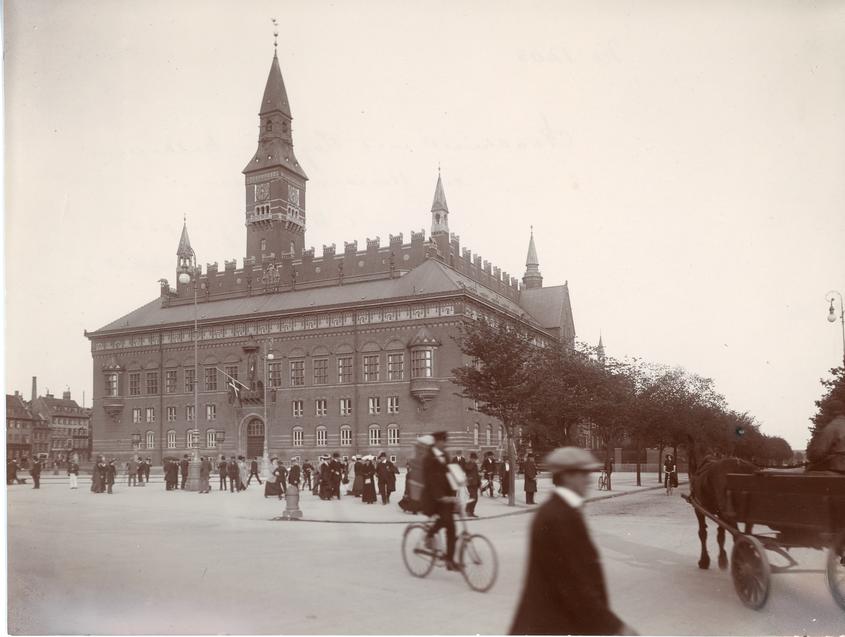 Gammelt billede af Københavns Rådhus i sepia-toner. Billedet er taget fra hjørnet af Rådhuspladsen ud mod Dagmarhus, og der passerer personer forbi - både gående, cyklende og i hestetrukne vogne. Billedet er taget i starten af 1900-tallet.